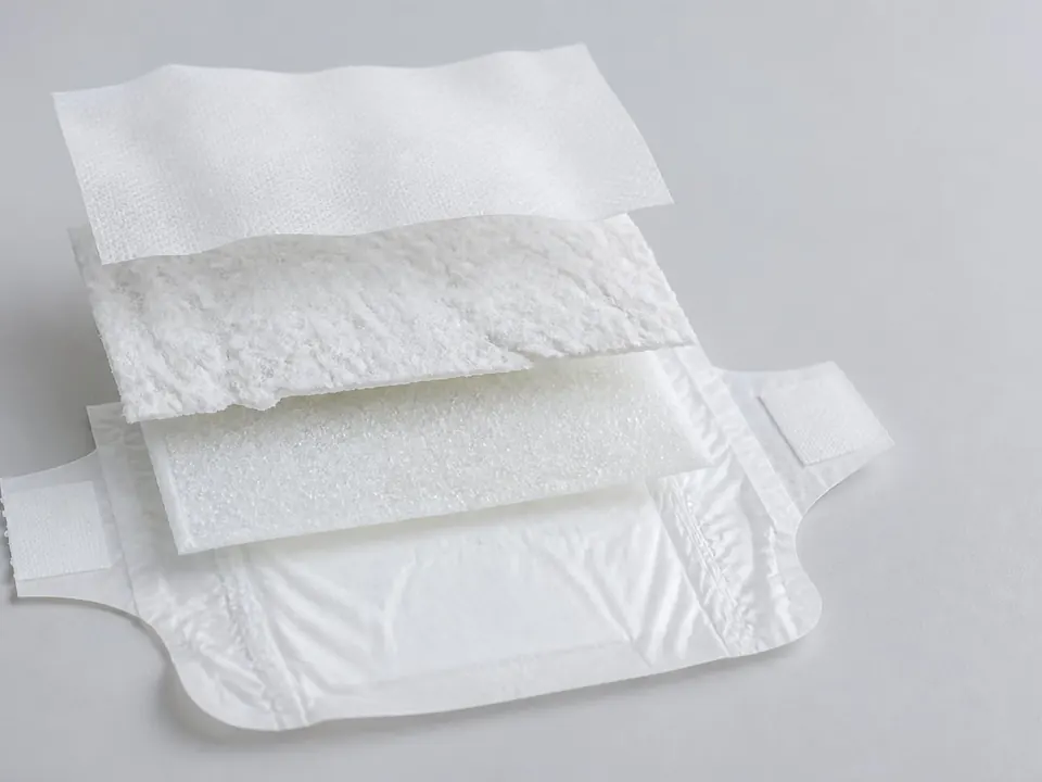 Opened disposable diaper in exploded view: inner liner, pulp, superabsorbent and plastic outer layer stacked as bonded layers.